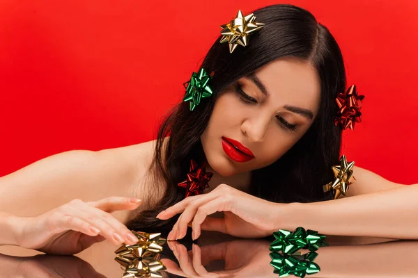 Jolie femme aux épaules nues et aux nœuds cadeaux brillants touchant une surface réfléchissante isolée sur rouge — Photo de stock