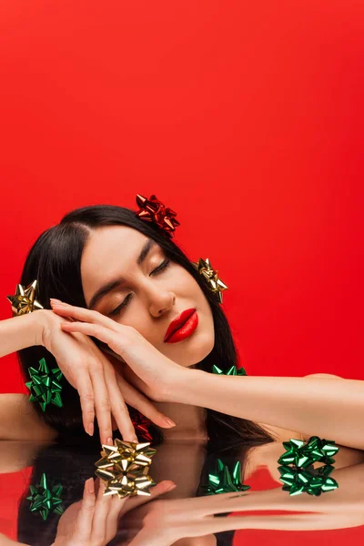 Jolie jeune modèle avec des noeuds cadeaux sur les cheveux posant avec les yeux fermés près de surface réfléchissante isolée sur rouge — Photo de stock