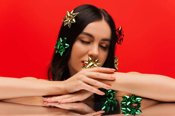 Jeune femme avec maquillage et noeuds cadeaux sur les cheveux toucher la main près de surface réfléchissante isolé sur rouge — Photo de stock