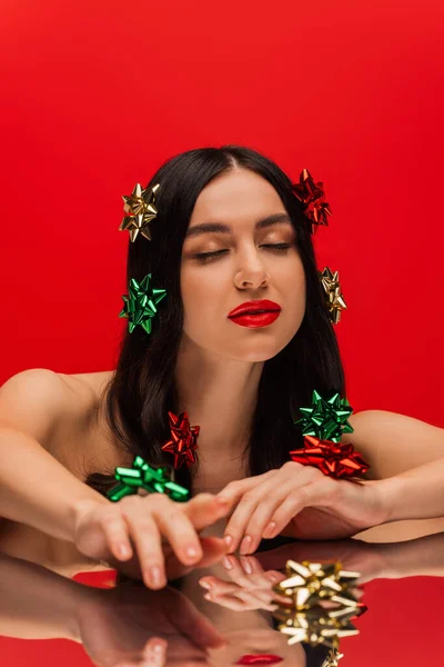 Jeune femme avec des noeuds cadeaux sur les cheveux pointant vers la caméra près de surface réfléchissante isolée sur rouge — Photo de stock