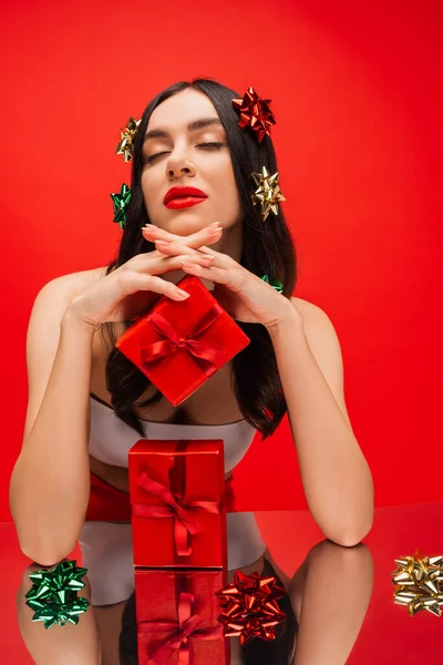 Brunette femme avec des noeuds cadeaux sur les cheveux posant près de cadeaux et surface réfléchissante isolée sur rouge — Photo de stock