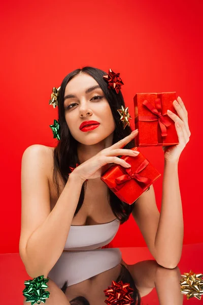 Jeune modèle en haut et noeuds cadeaux sur la tenue de cheveux présente près de surface réfléchissante isolé sur rouge — Photo de stock