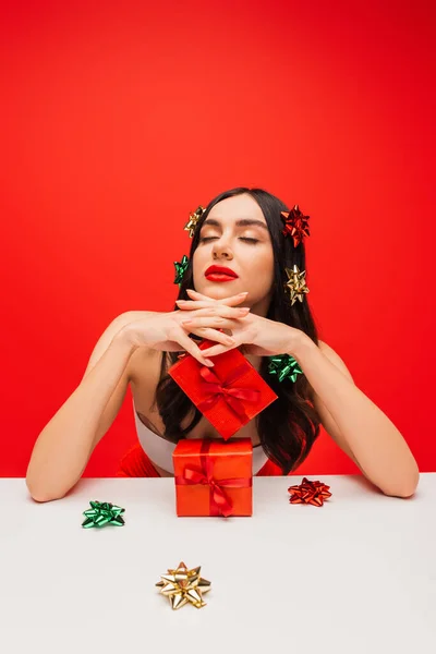 Красива жінка зверху і подарункові луки на волоссі позує біля подарунків ізольовані на червоному — стокове фото