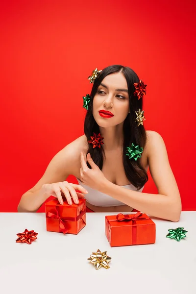 Jolie jeune femme avec des noeuds cadeaux sur les cheveux regardant loin près de cadeaux isolés sur rouge — Photo de stock