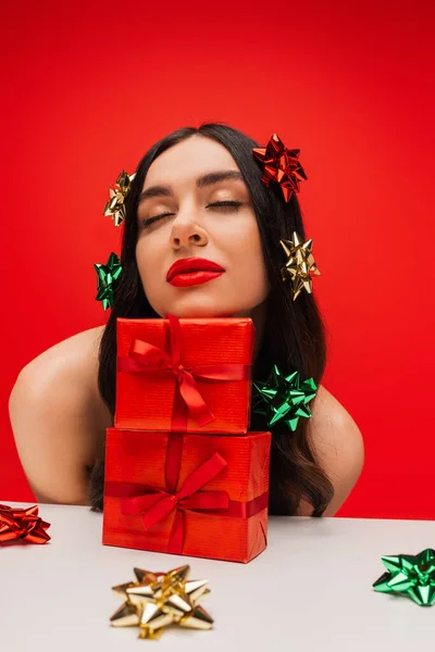 Modelo morena con brillantes lazos de regalo en el cabello posando cerca de regalos aislados en rojo - foto de stock