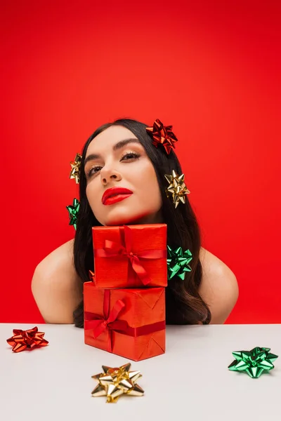 Jeune modèle avec des noeuds cadeaux sur les cheveux regardant loin près de cadeaux isolés sur rouge — Photo de stock