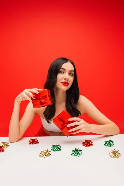Pretty model with visage holding presents near gift bows isolated on red — Stock Photo