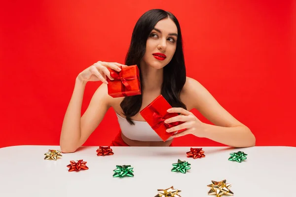 Femme brune avec maquillage tenant des cadeaux près de noeuds cadeaux brillants isolés sur rouge — Photo de stock
