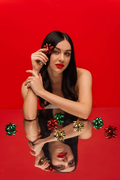 Brunette woman with naked shoulders holding shiny gift bows near reflective surface isolated on red — Stock Photo