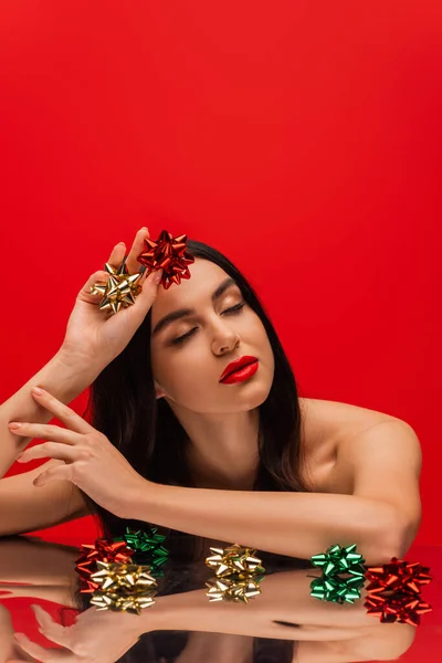 Hübsches Modell mit Make-up, das Geschenkschleifen hält, während es in der Nähe der reflektierenden Oberfläche posiert, isoliert auf Rot — Stockfoto