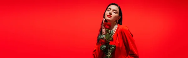 Модная брюнетка модель в куртке держа розы и закрывая глаза изолированы на красный, баннер — стоковое фото