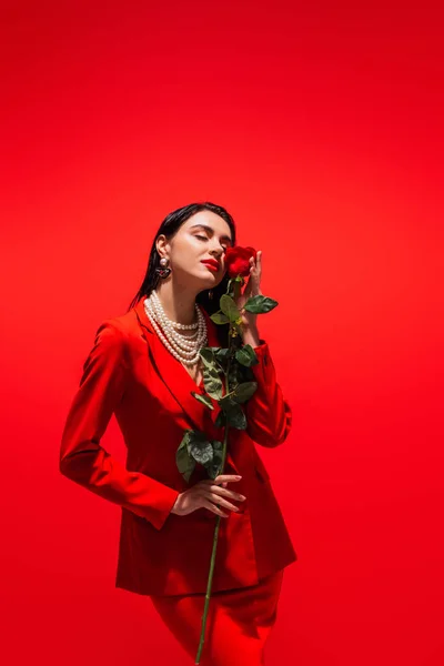 Стильна молода жінка в перловому намисті тримає троянду біля обличчя ізольовано на червоному — стокове фото