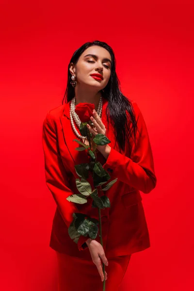 Fashionable woman in jacket holding rose while posing on red background — Stock Photo