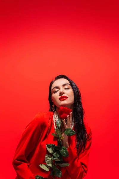 Elegant woman in pearl necklace holding rose and closing eyes isolated on red — Stock Photo