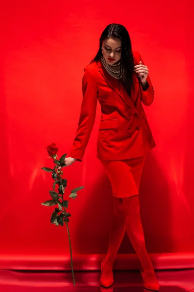 Longitud completa de la mujer con estilo en la chaqueta y collar de perlas sosteniendo rosa sobre fondo rojo - foto de stock