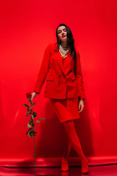 Volle Länge der trendigen brünetten Frau in Perlenkette mit Rose und Blick in die Kamera auf rotem Hintergrund — Stockfoto