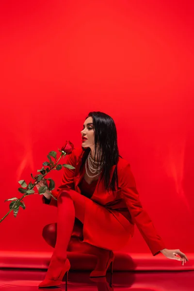 Стильная женщина в каблуках и ожерелье, держащая розу на красном фоне — стоковое фото