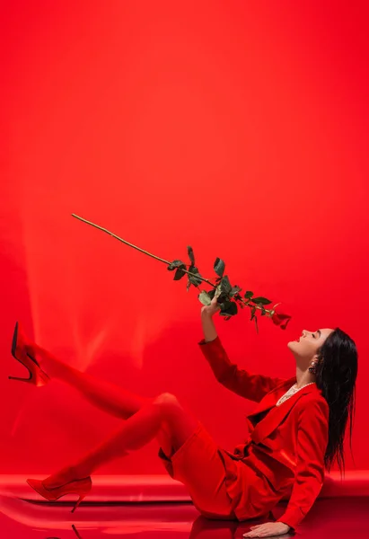 Seitenansicht des brünetten Modells in Heels, die Rose riechen, während sie auf rotem Hintergrund sitzen — Stockfoto