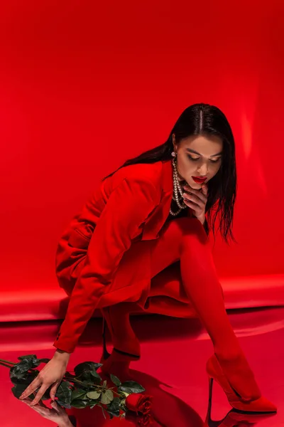 Stilvolles Modell in High Heels und Perlenkette auf reflektierender Oberfläche in der Nähe von Rose auf rotem Hintergrund — Stockfoto