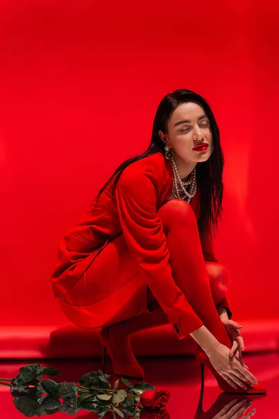 Stilvolle junge Frau in High Heels posiert in der Nähe von Rose auf reflektierender Oberfläche auf rotem Hintergrund — Stockfoto