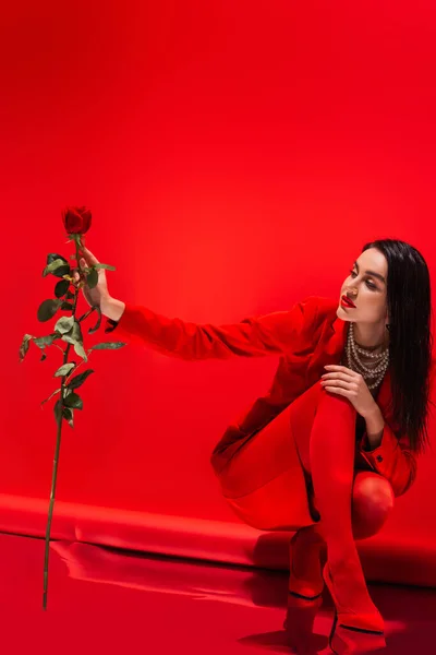 Стильна брюнетка в перловому намисті і на підборах дивиться на троянду на червоному тлі — стокове фото