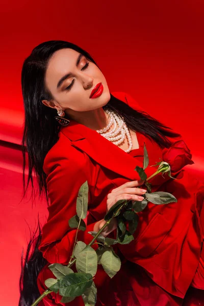 Junge Frau in Jacke und Perlenkette posiert mit Rose auf rotem Hintergrund — Stockfoto