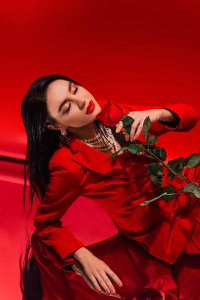 Молода жінка в куртці і перловому намисті тримає троянду і закриває очі на червоному тлі — стокове фото