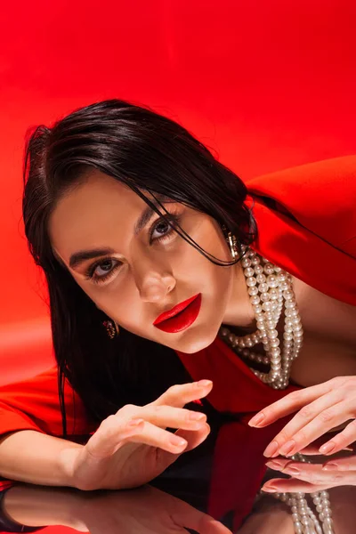 Элегантная женщина в жемчужном ожерелье смотрит на камеру рядом с отражающей поверхностью на красном фоне — стоковое фото