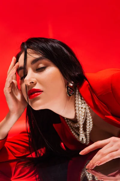 Retrato de mulher morena elegante no colar de pérolas deitado na superfície reflexiva isolada no vermelho — Fotografia de Stock