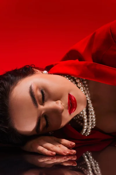 Jovem mulher em jaqueta vermelha e colar de pérolas deitado na superfície reflexiva no fundo vermelho — Fotografia de Stock