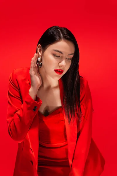 Elegant model in jacket touching earring while posing isolated on red — Stock Photo