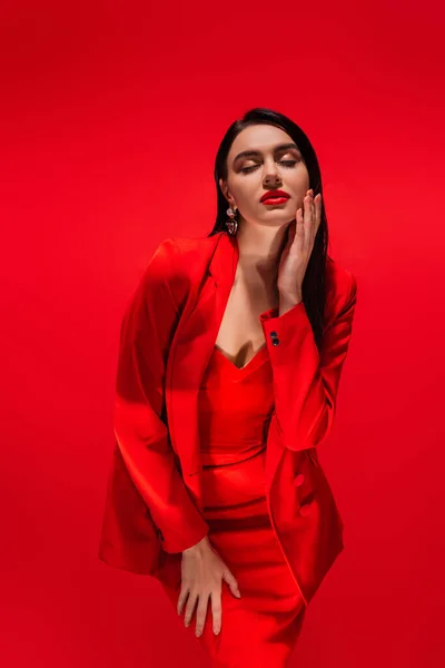 Modelo elegante e elegante na jaqueta tocando o rosto e fechando os olhos isolados no vermelho — Fotografia de Stock