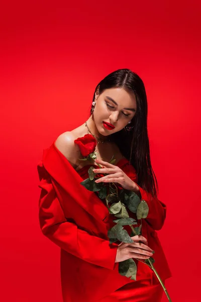 Stylish brunette model in jacket holding rose while closing eyes isolated on red — Stock Photo