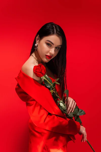 Стильная брюнетка, держащая розу у голого плеча на красном фоне — стоковое фото