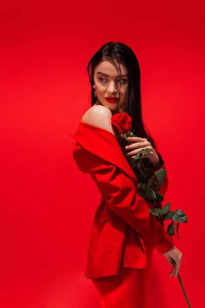 Elegant brunette woman in jacket with naked shoulder holding rose isolated on red — Stock Photo