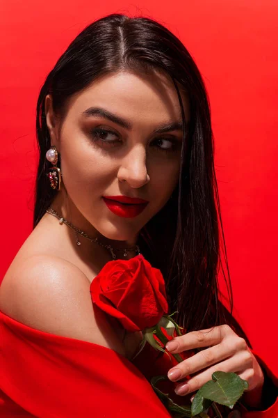 Porträt einer jungen brünetten Frau mit Visage, die Rosenblume in der Hand hält und isoliert auf Rot wegschaut — Stockfoto