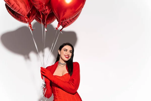 Mujer sonriente con labios rojos sosteniendo globos en forma de corazón sobre fondo blanco - foto de stock