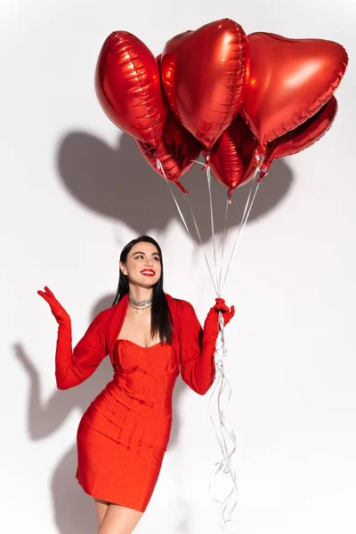 Positive und stilvolle Frau in roter Kleidung mit Blick auf herzförmige Luftballons auf weißem Hintergrund mit Schatten — Stockfoto
