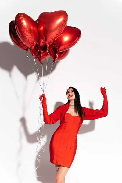 Весела брюнетка в червоній сукні дивиться на повітряні кулі у формі серця на білому тлі з тіні — стокове фото