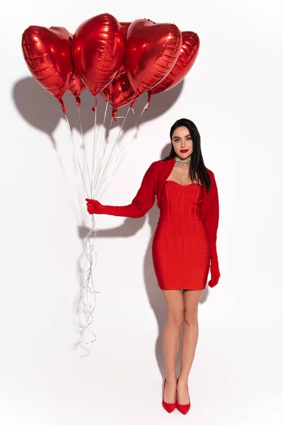 Longitud completa de la mujer con estilo en vestido rojo y tacones sosteniendo globos en forma de corazón sobre fondo blanco con sombra - foto de stock