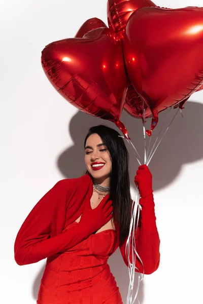 Mulher morena positiva em vestido vermelho posando com balões em forma de coração no fundo branco com sombra — Fotografia de Stock