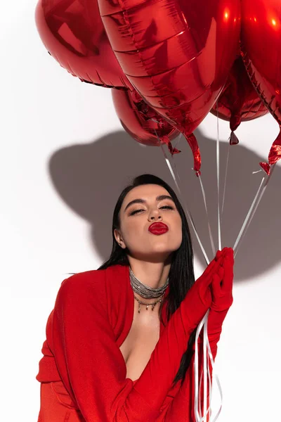 Mulher morena elegante em luvas vermelhas segurando balões em forma de coração e soprando beijo de ar no fundo branco com sombra — Fotografia de Stock