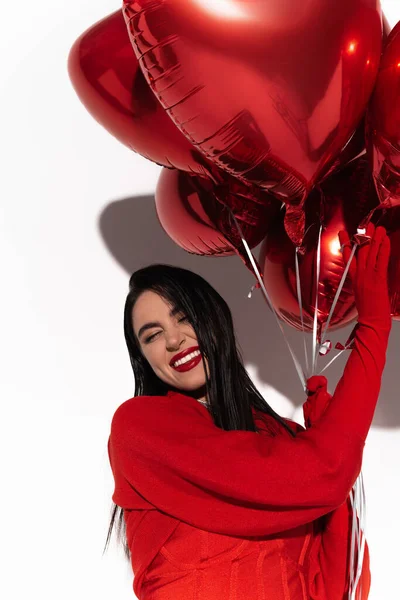Glückliche brünette Frau in roter Jacke, die herzförmige Luftballons auf weißem Hintergrund mit Schatten berührt — Stockfoto