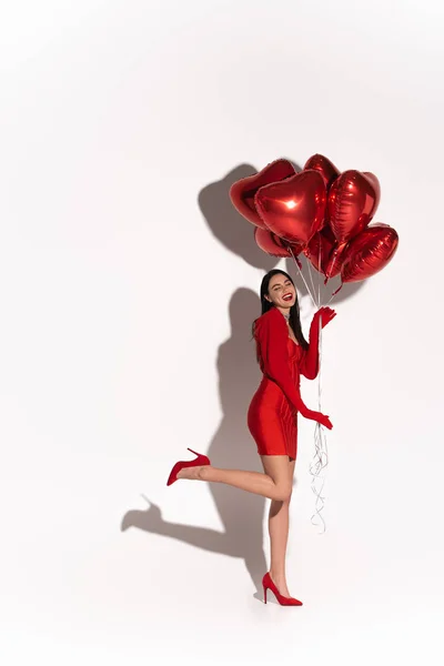 Pleine longueur de jolie femme en talons rouges et robe posant avec des ballons en forme de coeur sur fond blanc avec ombre — Photo de stock