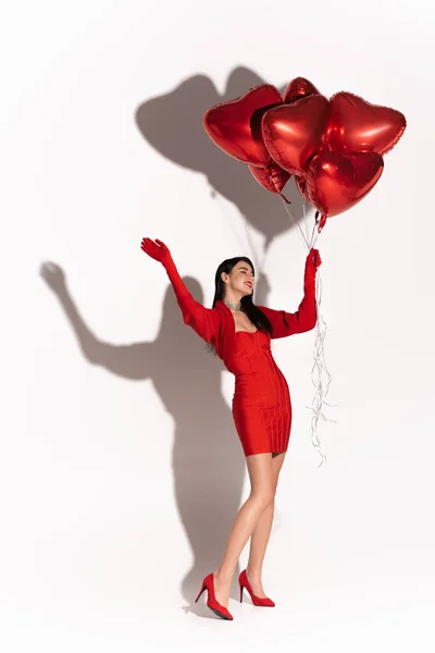 Cheerful woman in heels and dress holding red heart shaped balloons and waving hand on white background with shadow — Stock Photo
