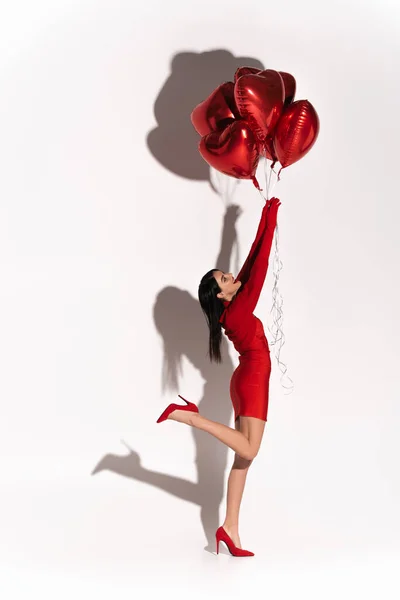 Возбужденная молодая женщина в красной одежде и на каблуках держит шары в форме сердца на белом фоне с тенью — стоковое фото