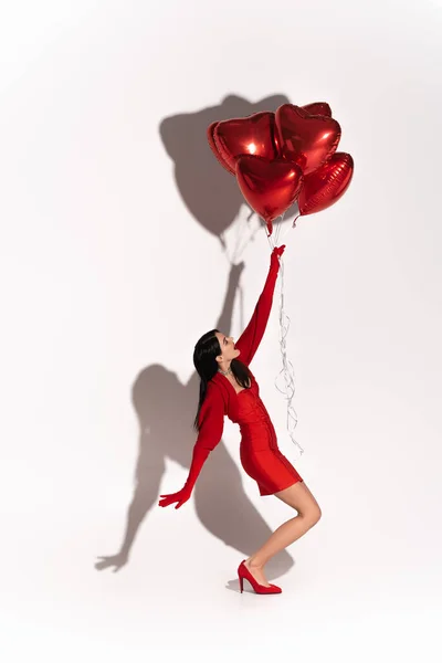 Vista lateral da mulher elegante no vestido e saltos puxando balões em forma de coração no fundo branco com sombra — Fotografia de Stock