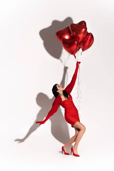 Vista lateral do modelo elegante no vestido olhando para balões em forma de coração no fundo branco com sombra — Fotografia de Stock