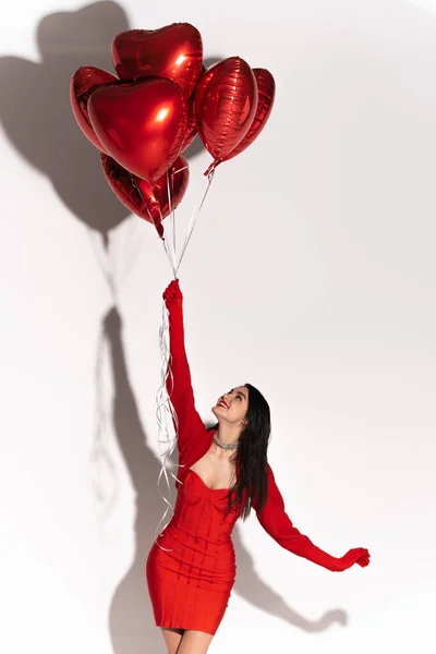 Mujer alegre en vestido y guantes mirando globos rojos en forma de corazón sobre fondo blanco con sombra - foto de stock