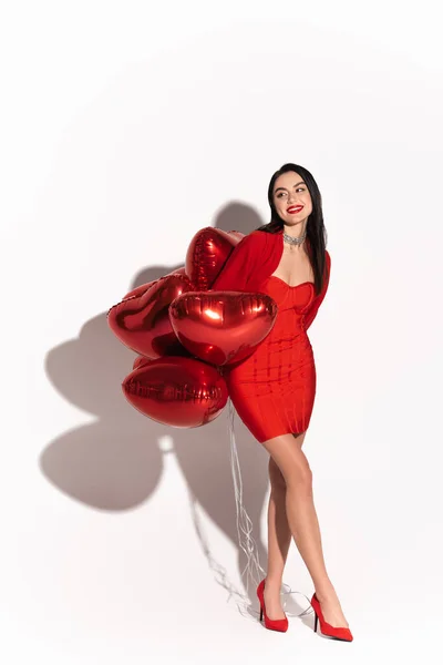 Стильная брюнетка женщина на каблуках глядя в сторону красных шаров в форме сердца на белом фоне с тенью — стоковое фото